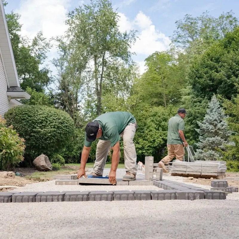 Two certified Techo-Pros installing a patio in a backyard surrounded by trees and shrubs, carefully laying pavers.