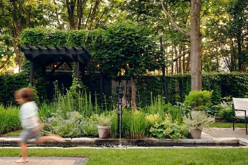 Family backyard garden with Limestone cap edging, water feature, pergola, lush planting, and a child running across the lawn.
