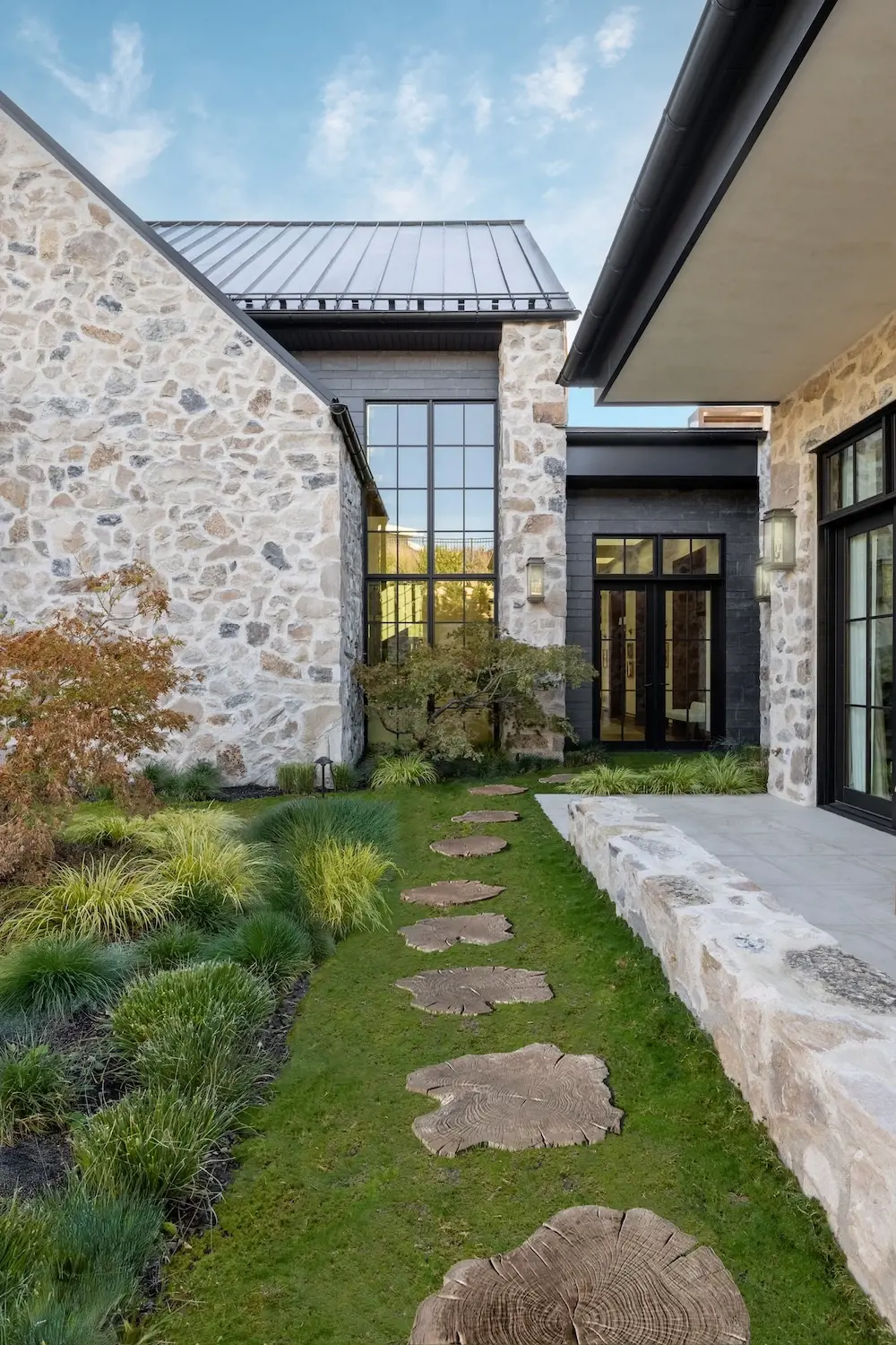 Courtyard walkway with large Maya stepping stones, lush garden beds, stone walls, and a modern glass entry.