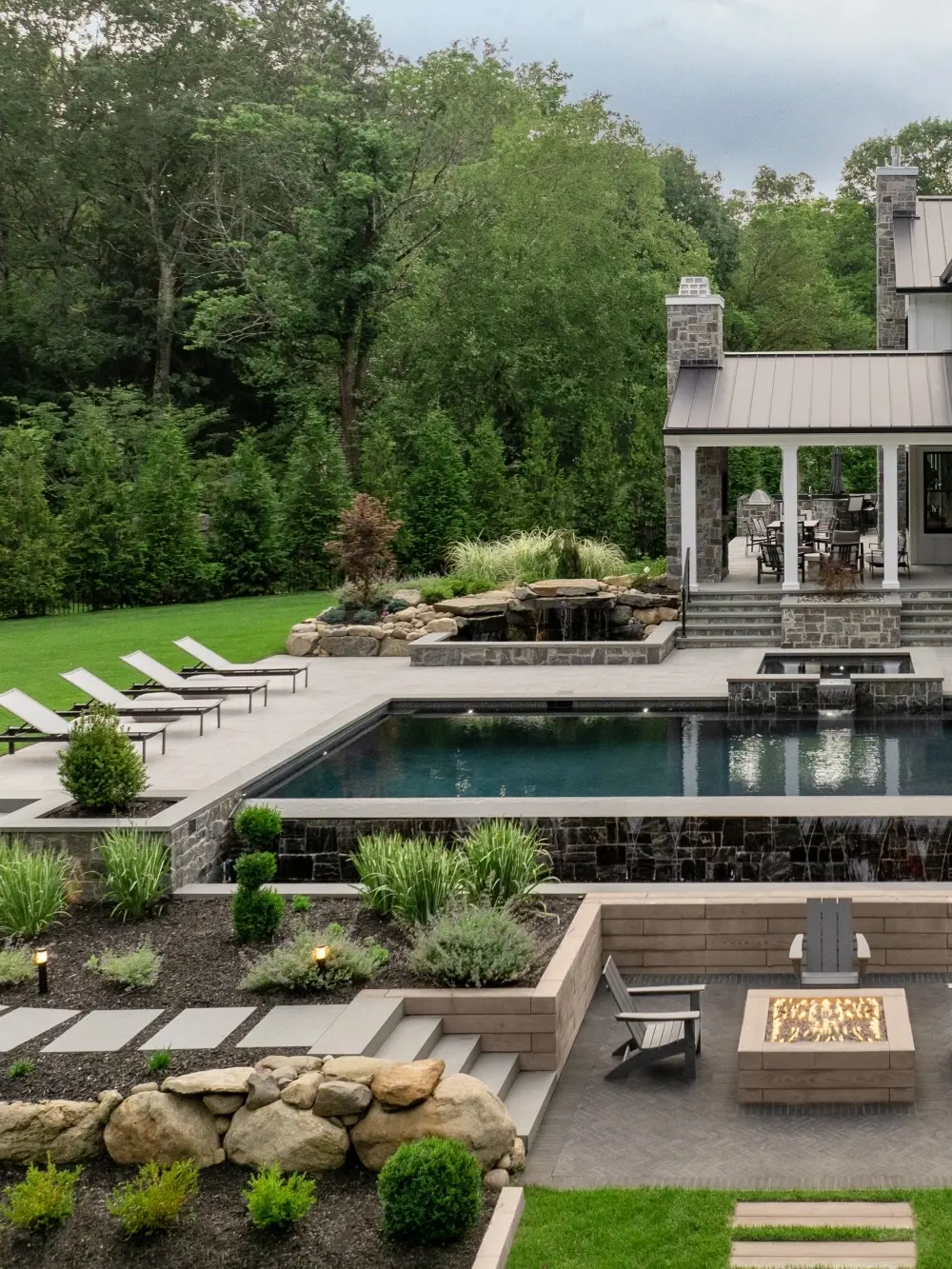 Cour arrière haut de gamme avec piscine, patio à niveaux multiples, murs de soutènement et foyer en pavés Techo-Bloc